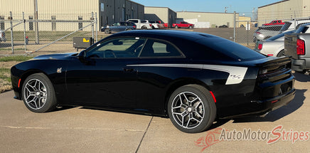 Black 2026 Dodge Charger Sixpack with side body vinyl graphics and stripes parked on a concrete surface with a building and other vehicles in the background.